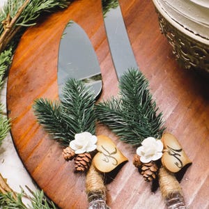 May include: A set of two silver cake knives with wooden heart-shaped handles decorated with pine sprigs, white flowers, and pine cones. The handles are tied with twine and have the initials "H" and "F" written on them.