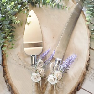 May include: A set of two silver cake cutting knives with rustic brown twine, white flowers, and purple lavender accents. The knives are on a wooden slice.
