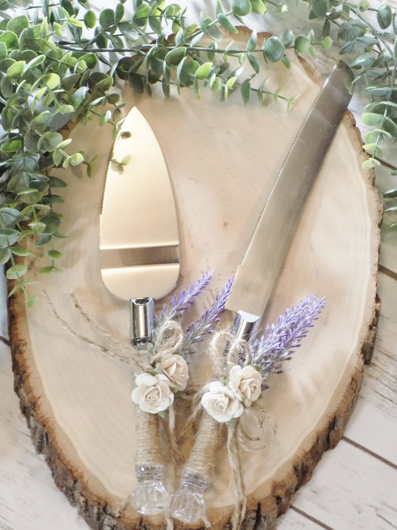 May include: A set of two silver cake cutting knives with rustic brown twine, white flowers, and purple lavender accents. The knives are on a wooden slice.
