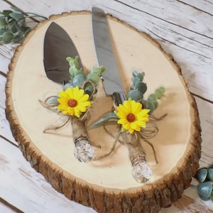 May include: A set of two cake cutting knives with rustic sunflower and eucalyptus accents. The knives are wrapped in burlap and tied with twine. The knives are displayed on a wooden slice.