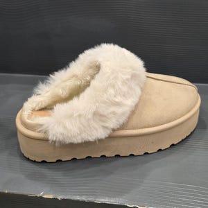 Puede incluir: Zapatilla de plataforma beige con un cuello esponjoso de color crema. La zapatilla tiene una puntera redondeada y una suela gruesa y texturizada. El interior parece estar forrado con un material suave. La zapatilla está diseñada para uso en interiores.