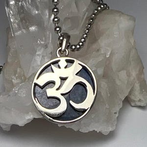 May include: A circular silver pendant featuring a cutout Om symbol set against a dark, textured background. The pendant hangs from a silver ball chain. The design is polished and metallic, showcasing a clear contrast between the silver symbol and the dark inlay within the circular frame, creating a distinct and recognizable spiritual emblem.