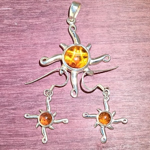 May include: A silver pendant and earrings set featuring amber stones in the shape of a sun. The pendant has a long, thin chain attached to it.