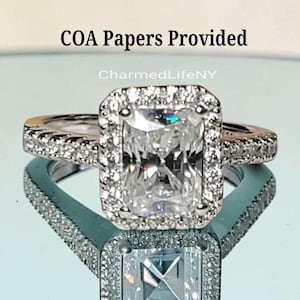 May include: A silver-toned ring featuring a large rectangular center stone surrounded by a halo of smaller clear stones. The band is also encrusted with small clear stones. Text above the ring reads COA Papers Provided and CharmedLifeNY.
