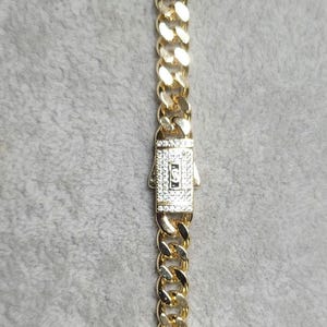 May include: A gold-toned cuban link chain necklace featuring a rectangular box clasp closure. The clasp is embellished with numerous small, clear, sparkling stones and displays a central rectangular emblem with the letter S engraved inside.