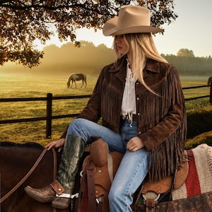 May include: A beige cowboy hat, a dark brown suede jacket with long fringe detailing, a white lace-up shirt, blue denim jeans, and tall green leather boots with metal spurs. The attire is worn while sitting on a brown leather saddle on a horse.