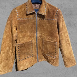 May include: A tan suede jacket featuring a full-length front zipper, a pointed collar, and two front patch pockets. The garment is decorated with silver-toned metal studs along the collar, the upper chest, and the edges of both pockets.
