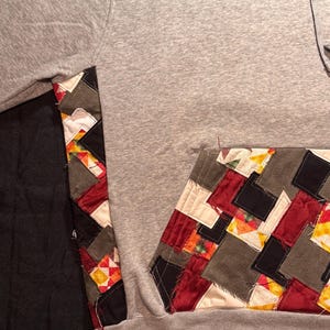 May include: A gray hoodie with a patchwork design on the pocket and sleeve. The patchwork features squares of red, black, gray, and yellow fabrics. The hoodie has a drawstring and a kangaroo pocket.
