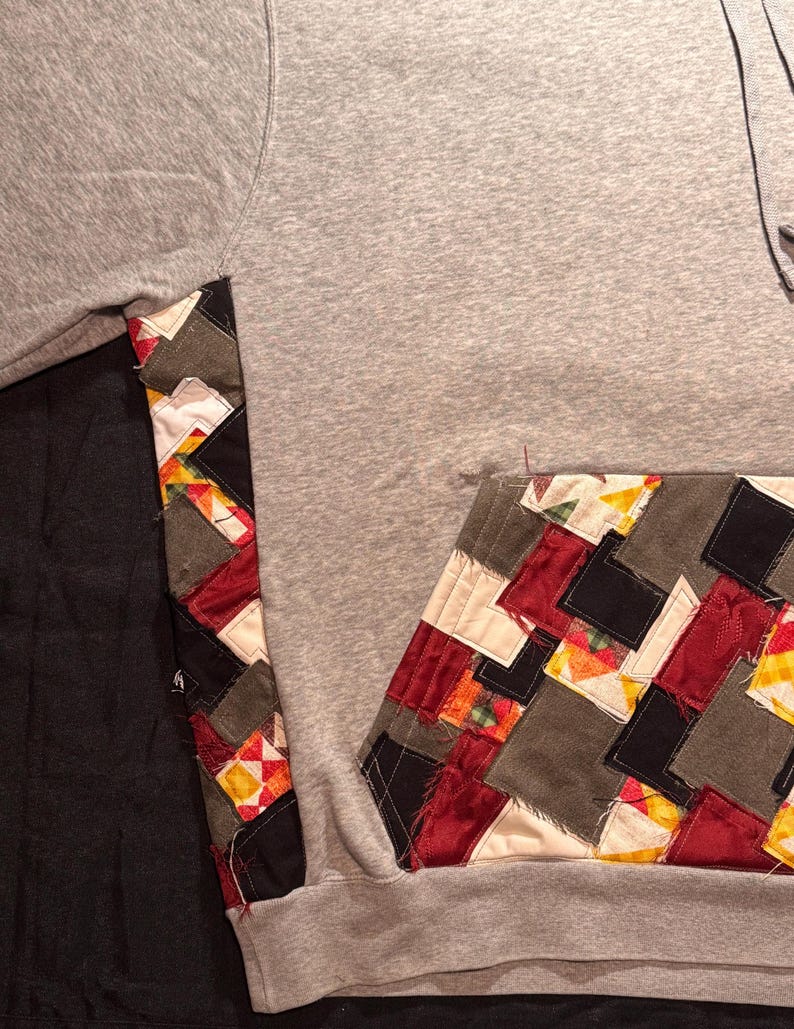 May include: A gray hoodie with a patchwork design on the pocket and sleeve. The patchwork features squares of red, black, gray, and yellow fabrics. The hoodie has a drawstring and a kangaroo pocket.