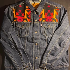 May include: A dark wash denim jacket with a colorful geometric design on the yoke. The design features red, orange, and yellow shapes on a black background. The jacket has button closures, pockets, and a classic collar.
