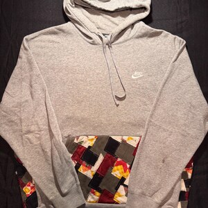 May include: A gray Nike hoodie with a patchwork pocket design. The hoodie features a drawstring hood and the Nike logo on the chest. The patchwork pocket includes squares of various colors and patterns.