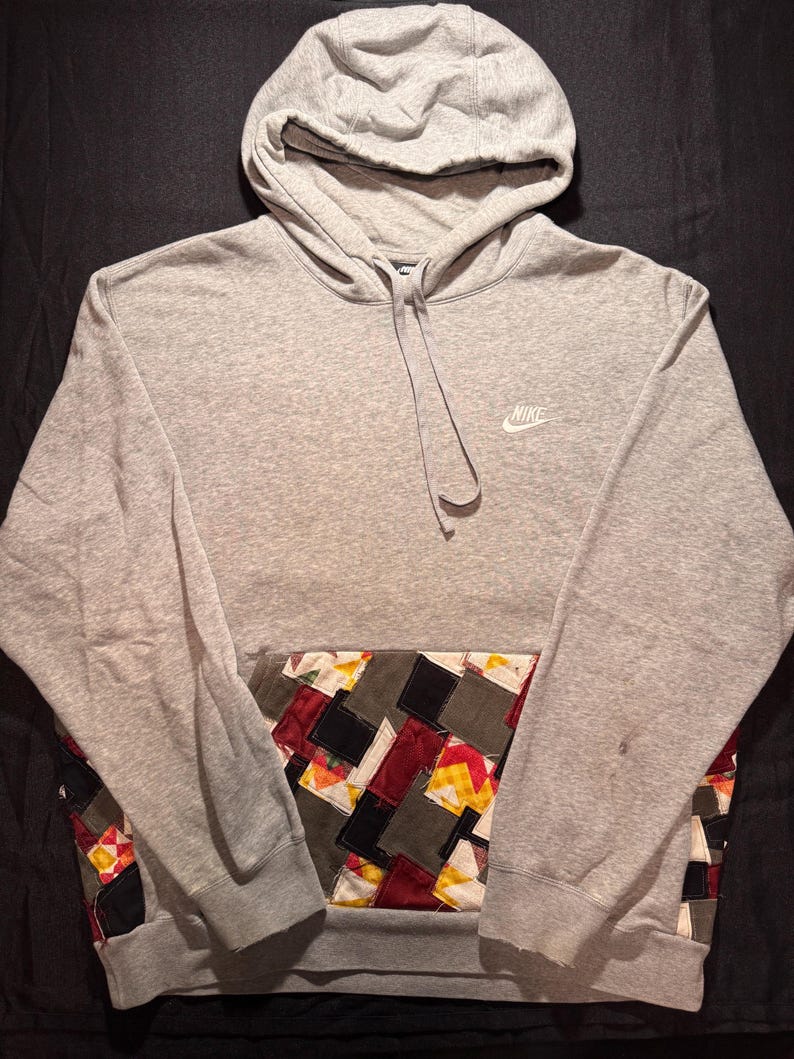 May include: A gray Nike hoodie with a patchwork pocket design. The hoodie features a drawstring hood and the Nike logo on the chest. The patchwork pocket includes squares of various colors and patterns.