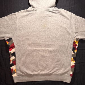 May include: A gray hooded sweatshirt with a patchwork design on the sides. The patchwork incorporates red, black, white, and yellow fabric pieces. The sweatshirt is made of a soft, comfortable material.