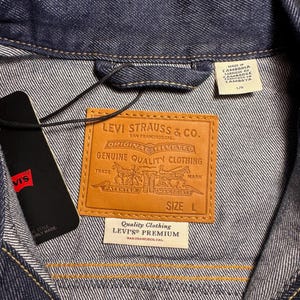 Puede incluir: Primer plano de una chaqueta vaquera azul oscuro con una etiqueta de cuero marr&oacute;n de Levi Strauss & Co. La etiqueta dice "ORIGINAL RIVETED GENUINE QUALITY CLOTHING". Una etiqueta negra con el logotipo de Levi's est&aacute; adjunta. La chaqueta es talla L.