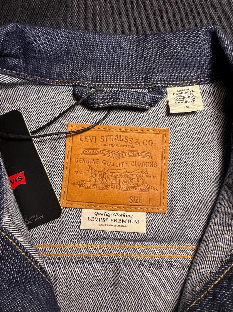 Puede incluir: Primer plano de una chaqueta vaquera azul oscuro con una etiqueta de cuero marr&oacute;n de Levi Strauss & Co. La etiqueta dice "ORIGINAL RIVETED GENUINE QUALITY CLOTHING". Una etiqueta negra con el logotipo de Levi's est&aacute; adjunta. La chaqueta es talla L.