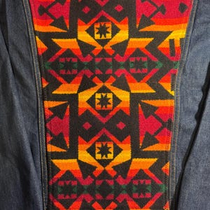 Puede incluir: Chaqueta vaquera azul con un llamativo panel trasero estampado. El panel presenta un dise&ntilde;o geom&eacute;trico repetitivo en tonos rojos, naranjas, amarillos y negros. La chaqueta es de estilo vaquero cl&aacute;sico.