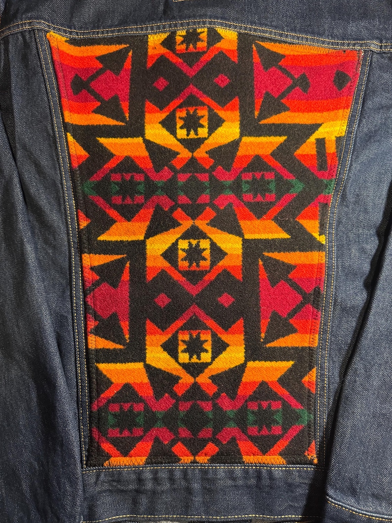 Puede incluir: Chaqueta vaquera azul con un llamativo panel trasero estampado. El panel presenta un dise&ntilde;o geom&eacute;trico repetitivo en tonos rojos, naranjas, amarillos y negros. La chaqueta es de estilo vaquero cl&aacute;sico.