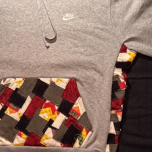 May include: A gray Nike hoodie with a patchwork pocket and sleeve detail. The patchwork incorporates various colors and patterns, including red, yellow, black, and gray. The Nike logo is visible on the chest.