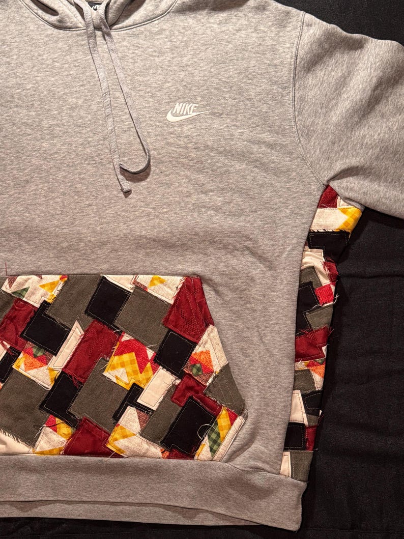 May include: A gray Nike hoodie with a patchwork pocket and sleeve detail. The patchwork incorporates various colors and patterns, including red, yellow, black, and gray. The Nike logo is visible on the chest.