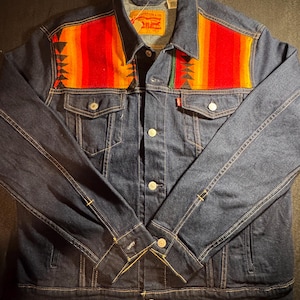 May include: A dark blue denim jacket with a colourful patterned yoke featuring red, orange, and yellow stripes. The jacket has button closures, front pockets, and yellow stitching. A small red tag is visible on the front.