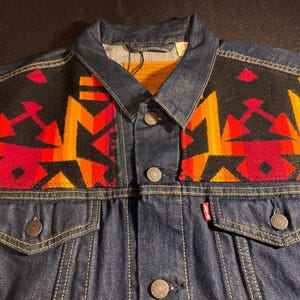 Puede incluir: Chaqueta vaquera azul oscuro con un colorido estampado geom&eacute;trico en los hombros. El estampado presenta formas rojas, naranjas y amarillas sobre un fondo negro. La chaqueta tiene botones y bolsillos.