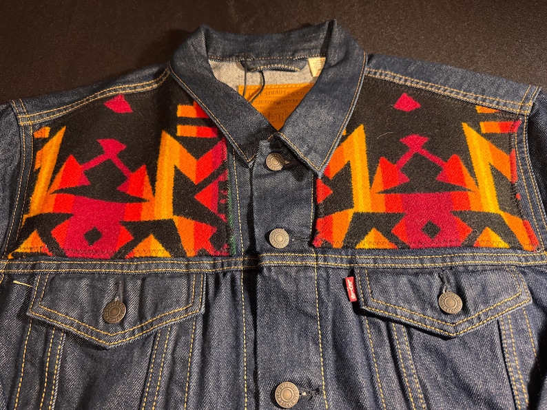 Puede incluir: Chaqueta vaquera azul oscuro con un colorido estampado geom&eacute;trico en los hombros. El estampado presenta formas rojas, naranjas y amarillas sobre un fondo negro. La chaqueta tiene botones y bolsillos.