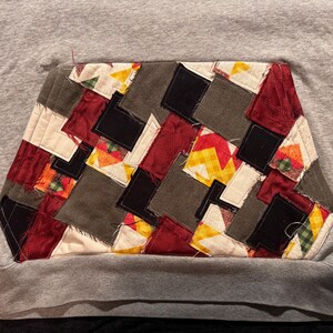 May include: A gray sweatshirt with a patchwork pocket. The pocket is made of various fabric squares in shades of red, black, gray, yellow, and white. The patchwork is quilted with white thread.