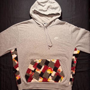 Puede incluir: Sudadera con capucha gris con un bolsillo y paneles laterales de patchwork. La sudadera tiene una capucha con cordón y el logotipo de Nike en blanco en el pecho. El diseño de patchwork incorpora varios colores y patrones.