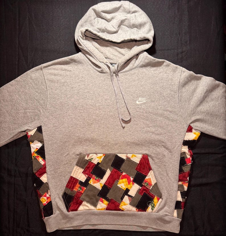 May include: Gray hoodie with a patchwork pocket and side panels. The hoodie features a drawstring hood and the Nike logo in white on the chest. The patchwork design incorporates various colors and patterns.