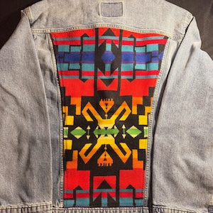 Op de afbeelding: Lichtblauwe spijkerjas met een opvallend, patroon achterpaneel. Het paneel toont een kleurrijk geometrisch ontwerp met rode, zwarte, blauwe, groene en gele vormen. De jas heeft een klassieke kraag, knoopsluitingen en zakken.