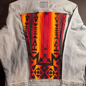 May include: Light blue denim jacket with a colorful patterned back panel. The panel features a geometric design in red, orange, yellow, green, and black. The jacket has a classic collar, button closures, and pockets.