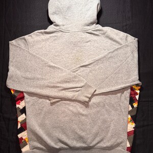 May include: A gray hoodie with a hood and long sleeves. The back of the hoodie features a colorful patchwork design along the lower sides. The hoodie is made of a soft, comfortable material.