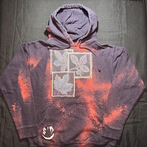Könnte beinhalten: Ein dunkelvioletter Kapuzenpullover mit einer Vordertasche. Das Design umfasst drei Blattmuster in quadratischen Feldern. Es hat einen roten Sprühfarbeffekt und einen kleinen Aufnäher mit einem lächelnden Gesicht und einem Dollarzeichen.