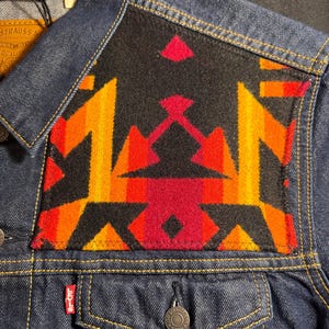Puede incluir: Chaqueta vaquera azul oscuro con un dise&ntilde;o geom&eacute;trico colorido en la parte superior de la espalda. El dise&ntilde;o presenta patrones rojos, naranjas y amarillos sobre un fondo negro. La chaqueta tiene una botonadura cl&aacute;sica y bolsillos.