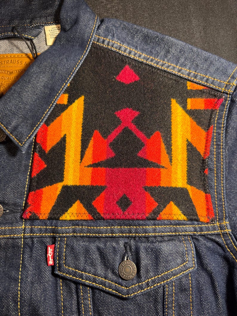 Puede incluir: Chaqueta vaquera azul oscuro con un dise&ntilde;o geom&eacute;trico colorido en la parte superior de la espalda. El dise&ntilde;o presenta patrones rojos, naranjas y amarillos sobre un fondo negro. La chaqueta tiene una botonadura cl&aacute;sica y bolsillos.