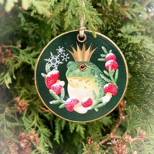Round wood slice ornament surrounded by evergreen foliage. The ornament is trimmed in gold, with a gold ribbon. Image is a green bullfrog wearing a gold crown, circled by a wreath of red mushrooms and spruce sprigs. Background is dark green.