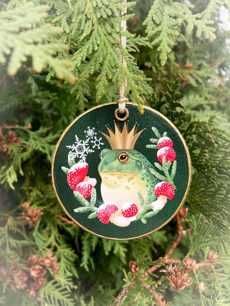 Round wood slice ornament surrounded by evergreen foliage. The ornament is trimmed in gold, with a gold ribbon. Image is a green bullfrog wearing a gold crown, circled by a wreath of red mushrooms and spruce sprigs. Background is dark green.