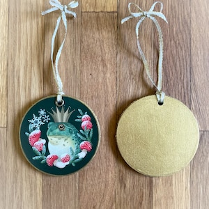 Wood circle Christmas ornament, shown front and back. Front image is a frog prince and Christmas wreath of forest flora in reds and greens, with metallic gold trim. Back is painted metallic gold. Ornament includes a gold ribbon.