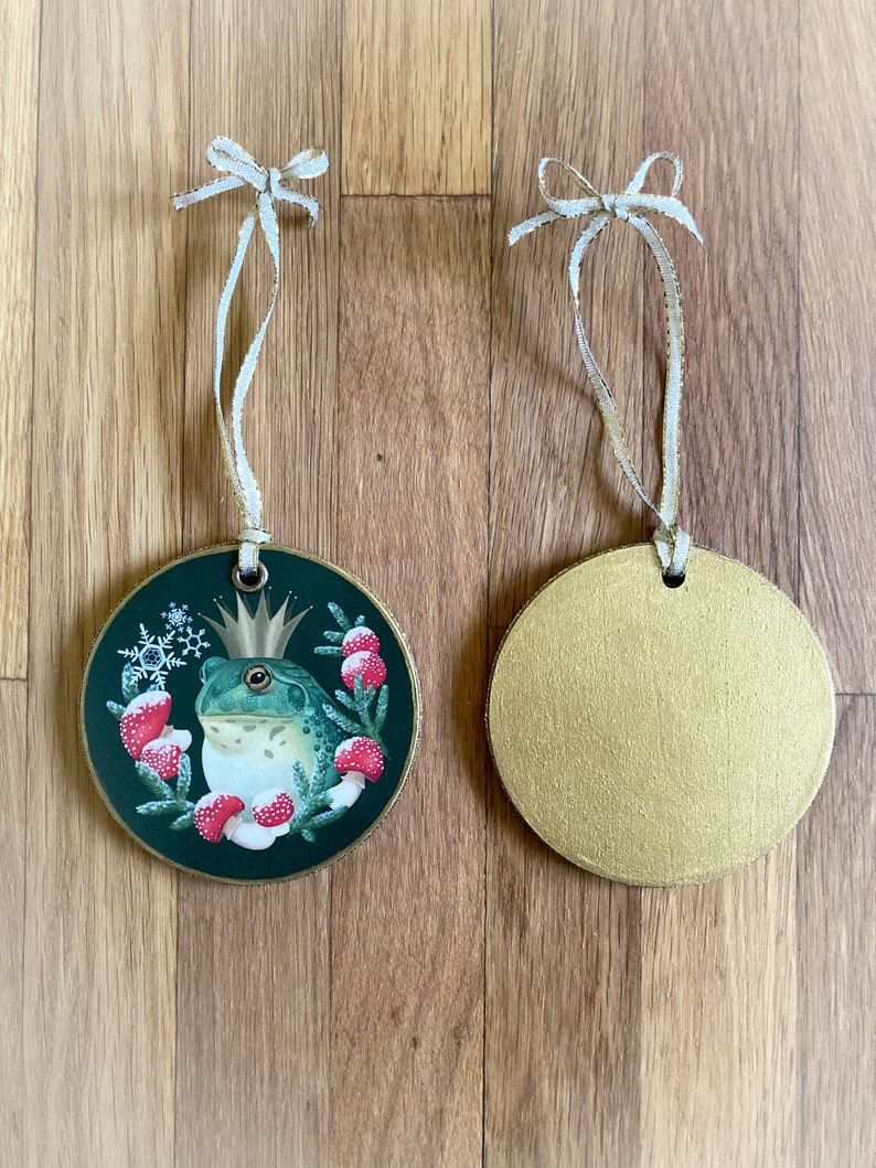 Wood circle Christmas ornament, shown front and back. Front image is a frog prince and Christmas wreath of forest flora in reds and greens, with metallic gold trim. Back is painted metallic gold. Ornament includes a gold ribbon.