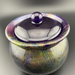 May include: A small, round, lidded container with a dark purple, glossy finish. The container has a spherical body with a lid and a small, round knob. The container is decorated with iridescent glitter that shifts colors.