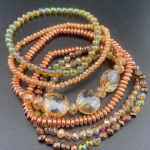 May include: A collection of beaded bracelets in shades of copper, gold, and iridescent colors. The bracelets feature various bead sizes and shapes, including small round beads and larger faceted beads. The bracelets are stacked together, creating a layered look.