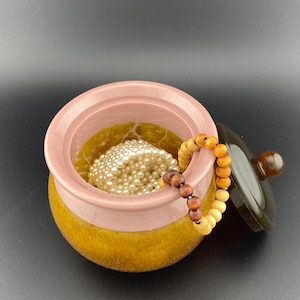 May include: A small, two-tone decorative pot with a lid. The pot is pink and gold, with a dark brown lid and a wooden bead bracelet draped over the rim. Inside the pot are white pearls.
