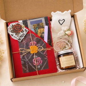 May include: A gift box filled with a red journal, a white sock with a heart, a candle, and a bracelet. The journal is decorated with a rose sticker and tied with twine. The candle has a label that reads "Aroma Sensations".