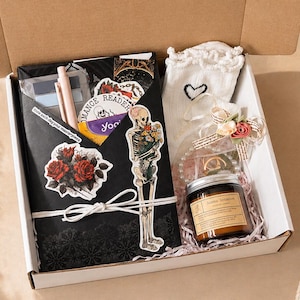 May include: An open gift box filled with various items. Includes a black envelope with stickers, a pen, and a small eyeshadow palette. Also, a pair of white socks with a heart, a candle, and a skeleton sticker.