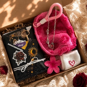 May include: A gift box containing a black notebook with celestial and rose stickers, a gold-tone key, a bright pink fuzzy pouch, a silver-tone necklace with a heart pendant, a pink flower-shaped accessory, and white socks with a red heart.
