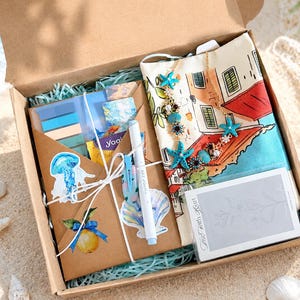 May include: A cardboard gift box containing a brown envelope with a lemon and jellyfish sticker, a light blue pen, a white fabric pouch with a Mediterranean building illustration, a blue starfish necklace, and a small card labeled The Earth Heart.