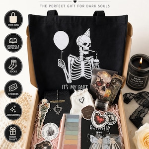 May include: A goth-themed gift box with a black tote bag featuring a skeleton holding a balloon. The box contains a journal, necklace, socks, stickers, accessories, and a candle. The box is filled with pink paper and includes a butterfly-themed item.