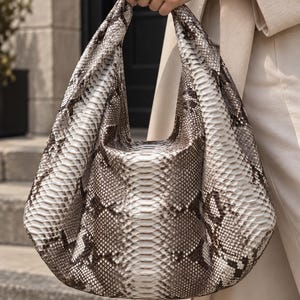 May include: A hobo-style handbag featuring a textured snakeskin pattern in shades of white, gray, and dark brown. The bag has a slouchy, unstructured silhouette with a single integrated handle, showcasing a detailed scale-like surface texture.