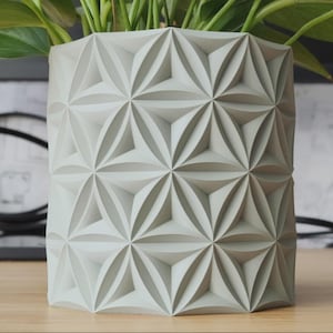 Könnte beinhalten: Ein zylindrischer, hellgrauer Übertopf mit einem sich wiederholenden geometrischen Muster aus vertieften Dreiecken und blütenblattartigen Formen. Die Oberfläche ist matt und weist scharfe, kantige Strukturen auf.