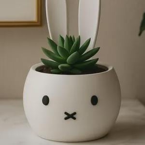 May include: A white ceramic planter shaped like a bunny face, with black eyes and a mouth. The planter holds a green succulent plant. The bunny ears are white and extend upwards. A decorative item for home decor.